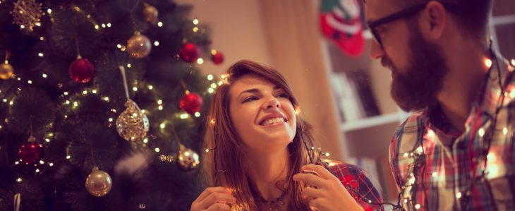 A man and a woman smile at each other while decorating a Christmas tree with a string of lights. 